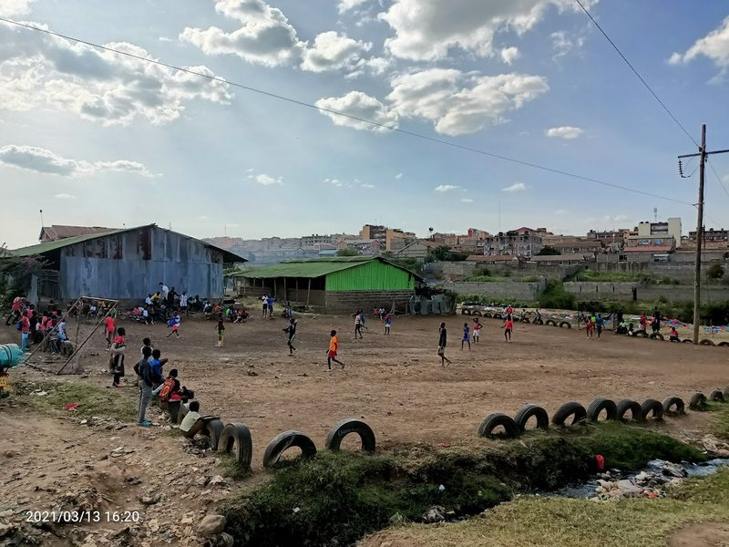 Terrain de football en terre délimité par des pneus, des enfants y jouent, Dandora en arrière-plan